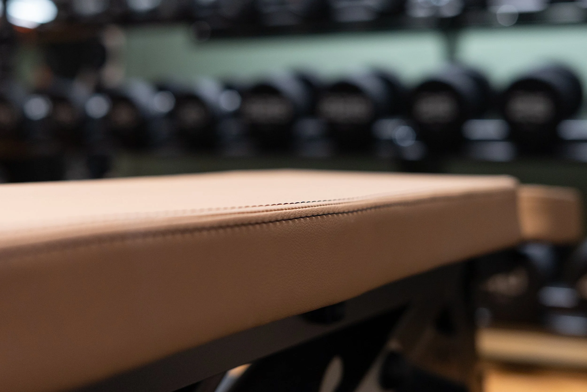 Close view of the stitched tan leather on a training bench, dumbbells out of focus behind